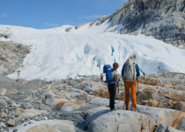 Two people are standing facing away from the camera. They are standing on rounded rocks in Greenland. The are looking towards some snow and ice.