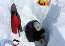 A person is sitting in a dug hole in the snow. A jacket is hung upon some equipment. A yellow box is seen on a shelf dug into the ice behind the person. The person is kneeling looking at something in their hands.