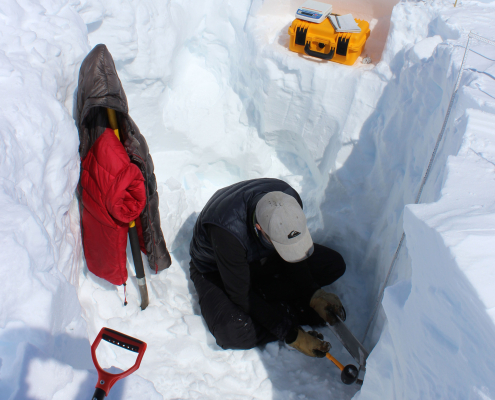 A person is sitting in a dug hole in the snow. A jacket is hung upon some equipment. A yellow box is seen on a shelf dug into the ice behind the person. The person is kneeling looking at something in their hands.