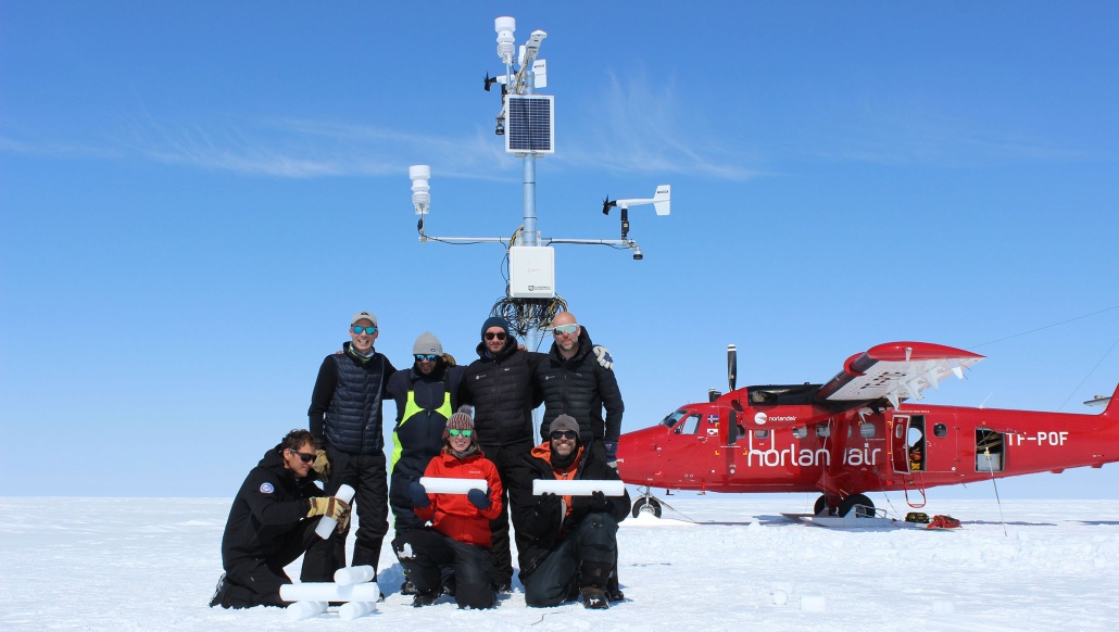 A group of people are standing and kneeling in the foreground, facing the camera. They are wearing fieldwork gear fit for work in the ice and snow. Behind them is a weather station. In the background is a red helicopter.